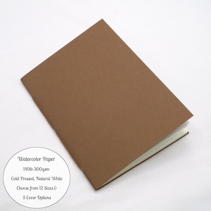 May include: A brown notebook with a textured cover. A circular white label features the text: Watercolor Paper, 140lb 300gsm, Cold Pressed, Natural White, Choose from 12 Sizes & 5 Cover Options. The notebook is shown at an angle.