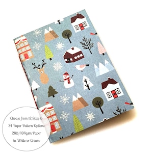 May include: A notebook with a winter-themed pattern featuring houses, trees, mountains, and a snowman on a light blue background. The text on the image says "Choose from 12 Sizes & 24 Paper Pattern Options 28lb/104gsm Paper in White or Cream."