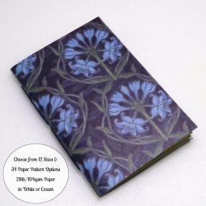 May include: A rectangular notebook with a dark purple cover featuring a repeating pattern of light blue flowers and green stems. Text on the cover reads: Choose from 12 Sizes & 34 Paper Pattern Options 28lb/104gsm Paper in White or Cream.