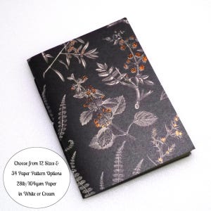 May include: A rectangular notebook with a black cover featuring a white and copper-colored botanical print of leaves and berries. Text on the cover reads: Choose from 12 Sizes & 34 Paper Pattern Options 28lb/104gsm Paper in White or Cream.