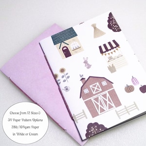 May include: A notebook with a white cover featuring a pattern of houses, a barn, a rabbit, sunflowers, pumpkins, and trees in shades of purple and brown. A light purple paper sheet is tucked behind the notebook. Text reads: Choose from 12 Sizes & 34 Paper Pattern Options 28lb/104gsm Paper in White or Cream.
