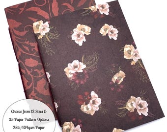 May include: Two notebooks with floral and patterned covers. One notebook has a dark burgundy cover with a repeating floral design. The other notebook has a dark brown cover with a floral pattern of white and burgundy flowers. The text on the image says "Choose from 12 Sizes & 35 Paper Pattern Options 28lb/104gsm Paper in White or Cream."