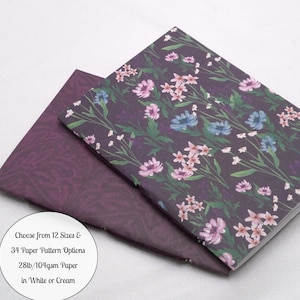 May include: Two notebooks with dark purple covers. The top notebook features a floral pattern with pink, blue, and white flowers on green stems. The bottom notebook has a dark purple tonal leaf pattern. Text reads: Choose from 12 Sizes & 34 Paper Pattern Options 28lb/104gsm Paper in White or Cream.