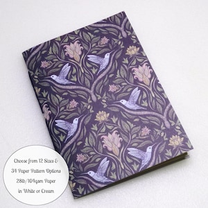 May include: A rectangular notebook featuring a dark purple cover with a repeating pattern of light purple hummingbirds, pale pink flowers, and green foliage. A circular white label includes the text: Choose from 12 Sizes & 34 Paper Pattern Options 28lb/104gsm Paper in White or Cream.