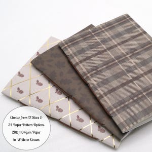 May include: Three notebooks with distinct covers: one with a gold grid and acorn pattern, one with a dark brown leaf print, and one with a brown plaid design. A circular label reads: Choose from 12 Sizes & 34 Paper Pattern Options 28lb/104gsm Paper in White or Cream.