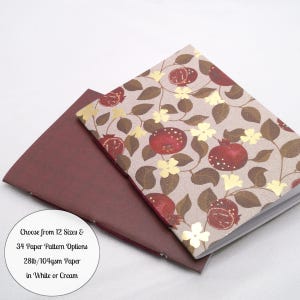 May include: Two notebooks with stitched spines. The top notebook features a light brown cover with a repeating pattern of red pomegranates, brown leaves, and gold flowers. The bottom notebook has a solid dark red cover. Text reads: Choose from 12 Sizes & 34 Paper Pattern Options 28lb/104gsm Paper in White or Cream.