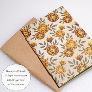 May include: A rectangular notebook with a floral pattern of yellow and orange marigolds on a light background, resting on a plain brown envelope. Text on the image reads: Choose from 12 Sizes & 34 Paper Pattern Options 28lb/104gsm Paper in White or Cream.