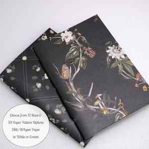 May include: Two notebooks with dark charcoal covers featuring botanical illustrations of white flowers, green leaves, and a small bird. A circular white label displays the text: Choose from 12 Sizes & 34 Paper Pattern Options 28lb/104gsm Paper in White or Cream.
