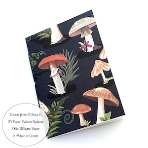 May include: A black notebook with a mushroom pattern. The mushrooms are red, brown, and white with green leaves. The text on the notebook says "Choose from 12 Sizes & 24 Paper Pattern Options 28lb/104gsm Paper in White or Cream".