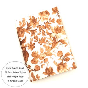 May include: A rectangular notebook with a white cover featuring a repeating copper-colored floral pattern. A circular text overlay states: Choose from 12 Sizes & 34 Paper Pattern Options 28lb/104gsm Paper in White or Cream.