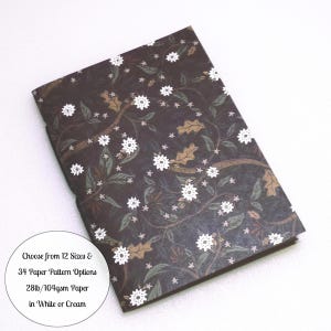 May include: A rectangular notebook featuring a dark brown cover with a repeating pattern of white flowers, green leaves, and tan oak leaves. A circular white label includes the text: Choose from 12 Sizes & 34 Paper Pattern Options 28lb/104gsm Paper in White or Cream.