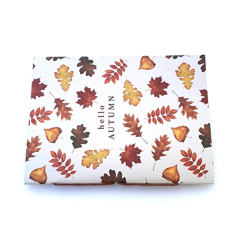 May include: A rectangular box with a white background, decorated with various autumn leaves in shades of brown, yellow, and orange. The text "hello AUTUMN" is printed vertically on the right side of the box.