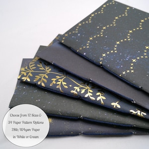 May include: A stack of five hand-bound notebooks with dark covers featuring gold foil patterns of stars and botanical vines. The spines are stitched with dark blue thread. Text on the image reads: Choose from 12 Sizes & 34 Paper Pattern Options 28lb/104gsm Paper in White or Cream.