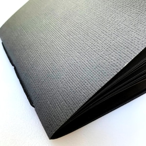 May include: A black notebook with a textured cover. The notebook is closed and the cover is slightly open, revealing the pages inside.