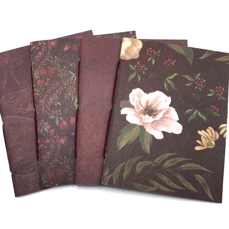 May include: A set of four rectangular paper notebooks featuring dark burgundy and brown covers. The designs include botanical illustrations of white flowers, green leaves, red berries, and moth outlines, all bound with thin dark red thread.