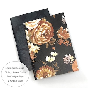May include: Two rectangular notebooks with black covers. One cover features a dark floral pattern with orange, yellow, and white flowers. The other has a subtle black-on-black damask print. Text reads: Choose from 12 Sizes & 34 Paper Pattern Options 28lb/104gsm Paper in White or Cream.