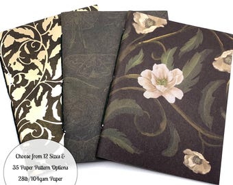 May include: Three rectangular notebooks with distinct covers: one with a cream and dark brown swirling vine pattern, one with a dark gray moth print, and one with a dark brown background featuring light pink flowers and green leaves. Text reads: Choose from 12 Sizes & 35 Paper Pattern Options 28lb/104gsm Paper in White or Cream.