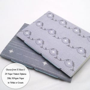 May include: Two notebooks with grey covers. The top notebook features a pattern of thin vertical lines and repeating vine motifs with stars. The bottom notebook has a solid grey cover with white star accents. Text reads: Choose from 12 Sizes & 34 Paper Pattern Options 28lb/104gsm Paper in White or Cream.