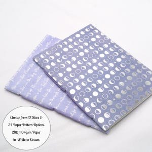 May include: Two notebooks with light purple covers. The top notebook features a repeating silver foil pattern of moon phases and small stars. The bottom notebook has white cursive text printed across the light purple surface.