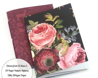 May include: Two notebooks with floral designs. The top notebook has a black cover with pink and red roses, while the bottom notebook has a burgundy cover with a deer pattern. The text on the bottom notebook reads "Choose from 12 Sizes & 24 Paper Pattern Options 28lb/104gsm Paper in White or Cream."