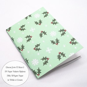 May include: A light green folded card featuring a repeating pattern of dark green holly leaves with red berries and white snowflakes. A circular text overlay states: Choose from 12 Sizes & 34 Paper Pattern Options 28lb/104gsm Paper in White or Cream.