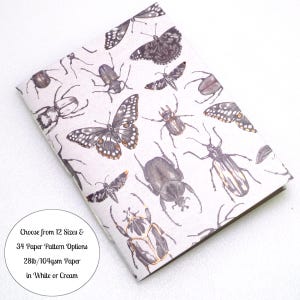 May include: A notebook with a white cover featuring a repeating pattern of various dark-colored beetles and butterflies. A circular white label on the cover includes the text: Choose from 12 Sizes & 34 Paper Pattern Options 28lb/104gsm Paper in White or Cream.