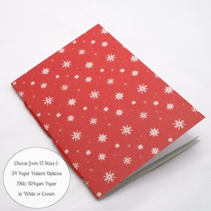 May include: A red notebook with a white snowflake pattern. A circular white label on the cover reads: Choose from 12 Sizes & 34 Paper Pattern Options 28lb/104gsm Paper in White or Cream. The notebook is shown at an angle with white pages visible.