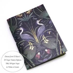 May include: A rectangular notebook featuring a dark purple and black botanical cover with illustrations of two herons, dragonflies, and cattails. Text on the cover reads: Choose from 12 Sizes & 34 Paper Pattern Options 28lb/104gsm Paper in White or Cream.