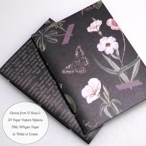 May include: Two rectangular notebooks with a dark charcoal background featuring a botanical print of light pink flowers, green leaves, and a butterfly. The cover includes purple tape accents and printed text. A circular white label displays text.