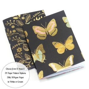 May include: Two notebooks with black covers. One features gold floral patterns, the other has gold butterflies with pink and teal accents. The text on the image says "Choose from 12 Sizes & 24 Paper Pattern Options 28lb/104gsm Paper in White or Cream."