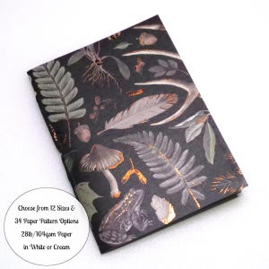 May include: A rectangular notebook with a black cover featuring a botanical and woodland pattern of ferns, mushrooms, feathers, acorns, and a frog, accented with copper foil details. Text reads: Choose from 12 Sizes & 34 Paper Pattern Options 28lb/104gsm Paper in White or Cream.