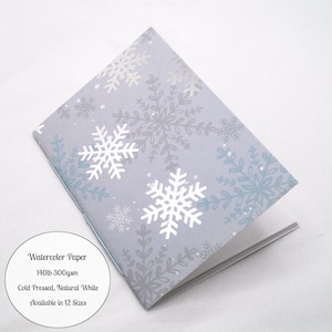 May include: A folded booklet with a light blue cover featuring a repeating pattern of white and silver snowflakes. A circular white label on the left displays the text: Watercolor Paper, 140lb 300gsm, Cold Pressed, Natural White, Available in 12 Sizes.