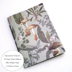 May include: A notebook with a light green cover featuring a botanical print of various mushrooms, ferns, and leaves. A circular white text overlay reads: Choose from 12 Sizes & 34 Paper Pattern Options 28lb/104gsm Paper in White or Cream.