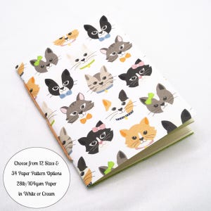 May include: A notebook with a white cover featuring a repeating pattern of various cat faces in black, gray, white, and orange, some wearing bows or collars. Text on the cover reads: Choose from 12 Sizes & 34 Paper Pattern Options 28lb/104gsm Paper in White or Cream.