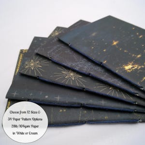 May include: A stack of five dark-colored notebooks with celestial star patterns in gold, bound with dark blue thread. A circular white label on the bottom left reads: Choose from 12 Sizes & 34 Paper Pattern Options 28lb/104gsm Paper in White or Cream.