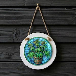 May include: Round white sign with a detailed illustration of cannabis plants in a pot, set against a blue stained-glass-style background. The sign hangs from a jute rope against a dark wood background.