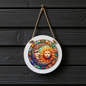 May include: Round white wall hanging featuring a colorful sun and moon design. The sun has a smiling face with orange and yellow rays, while the moon has a serene face with blue and purple accents. The design is surrounded by a decorative border.