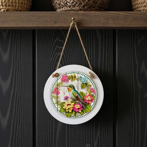 May include: Round white wall hanging with a hummingbird design. The artwork features a colorful hummingbird surrounded by pink, yellow, and blue flowers. The design is set against a white wooden background and framed with a green dotted border.