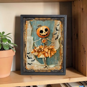 May include: A framed Halloween-themed artwork featuring a pumpkin head with a skeleton body and a skirt made of dried leaves. The artwork has a blue background with torn fabric and bat silhouettes. The frame is dark gray.