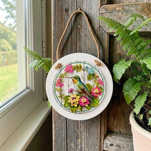 May include: A circular wall hanging featuring a central illustration of a hummingbird surrounded by a wreath of pink, yellow, and blue flowers. The artwork is set within a white frame and suspended by a brown twine rope for wall mounting.