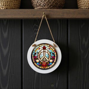 May include: Round white wall art with a stained glass design featuring a peace symbol, hands, and roses. The art is suspended by a natural rope and hangs against a dark wood background. The peace symbol is in the center.