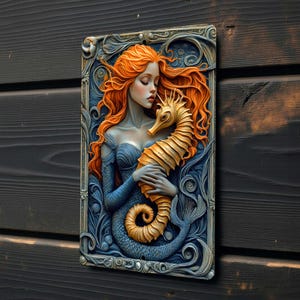 May include: A rectangular wall art piece featuring a mermaid with flowing orange hair embracing a golden seahorse. The mermaid is depicted in a blue dress with a blue tail. The background is a detailed, ornate frame with blue and silver accents.