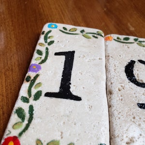 May include: Two white ceramic tiles with black painted numbers, 1 and 0, surrounded by a green vine with colorful flowers. The tiles are used for house numbers.
