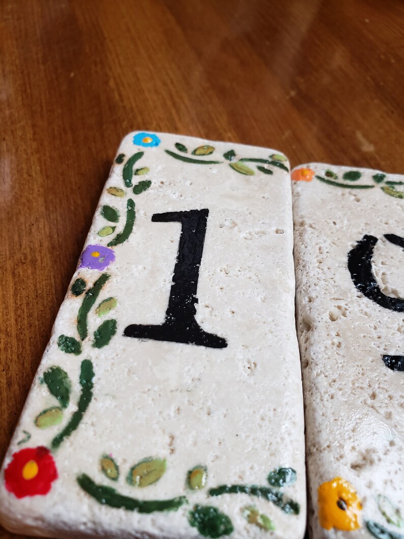 May include: Two white ceramic tiles with black painted numbers, 1 and 0, surrounded by a green vine with colorful flowers. The tiles are used for house numbers.