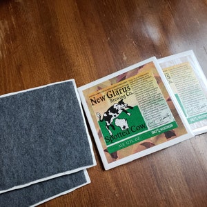 May include: Two grey coasters with a white border and a white square coaster with a green and brown illustration of a spotted cow and the text "New Glarus Brewing Co. Spotted Cow Ale 12 FL OZ. Only in Wisconsin."