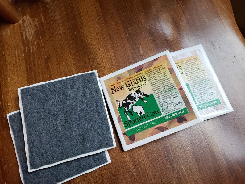 May include: Two grey coasters with a white border and a white square coaster with a green and brown illustration of a spotted cow and the text "New Glarus Brewing Co. Spotted Cow Ale 12 FL OZ. Only in Wisconsin."