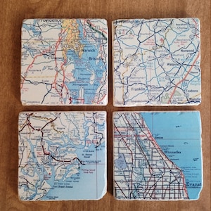 May include: Four square stone coasters featuring printed road map sections of various regions. Each coaster displays detailed cartographic information including city names, highway numbers, and water bodies in blue, red, and black ink on a white background.
