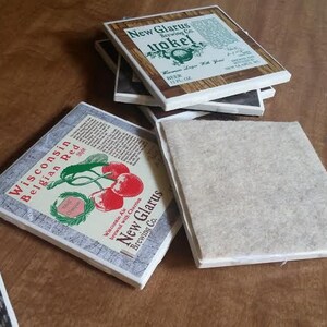May include: A stack of four square coasters with a brown wood grain design. The top coaster features a white background with the text "New Glarus Brewing Co." and "Yokel" in a brown font. The coaster below it has a white background with the text "Wisconsin Belgian Red" and a red cherry graphic. The bottom two coasters are plain brown.