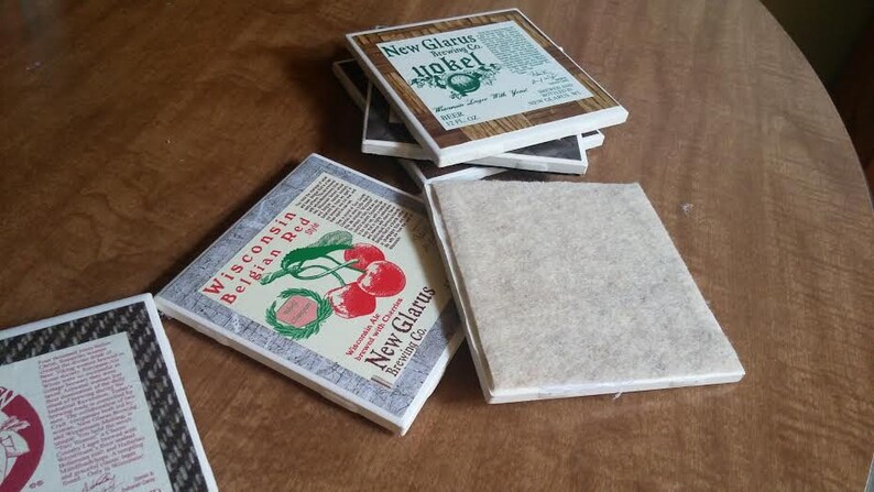 May include: A stack of four square coasters with a brown wood grain design. The top coaster features a white background with the text "New Glarus Brewing Co." and "Yokel" in a brown font. The coaster below it has a white background with the text "Wisconsin Belgian Red" and a red cherry graphic. The bottom two coasters are plain brown.