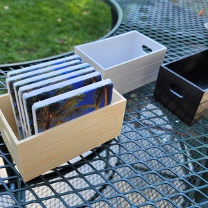 Puede incluir: Un juego de posavasos rectangulares con un dise&ntilde;o de palmera y cielo azul, exhibidos en una caja de madera clara. Tambi&eacute;n se ven dos cajas de madera adicionales, una blanca y otra negra.